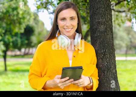 Lächelnde Frau in gelbem Hemd, die draußen im Park ein Tablet hält und in die Kamera schaut Stockfoto