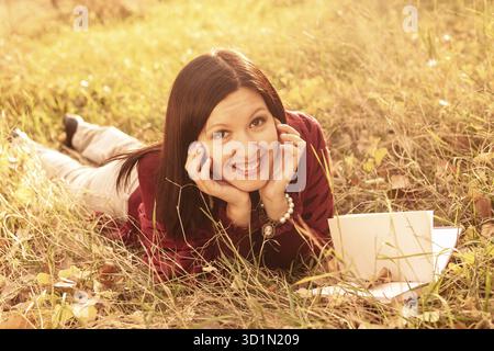 Glückliche Frau, die mit Buch auf dem herbstlichen Grasfeld liegt Stockfoto