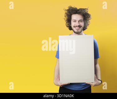 Bärtiger Mann mit Schnurrbart und lustigen Haaren, der ein leeres weißes Brett auf orangefarbenem Hintergrund hält Stockfoto