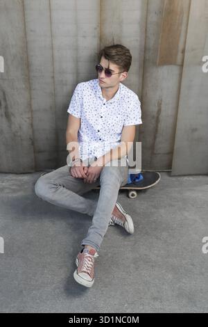 Junger, stilvoller Mann in Sonnenbrille, der auf einem Skateboard sitzt Stockfoto