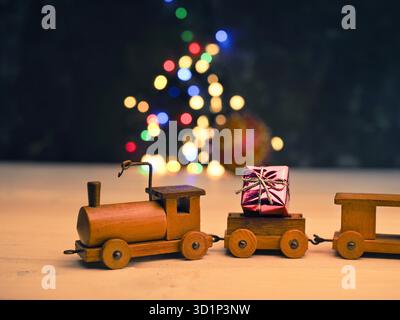 Kleiner hölzerner Zug mit Geschenkbox auf verschwommenen Weihnachtslichtern, saisonaler oder weihnachtlicher Hintergrund Stockfoto