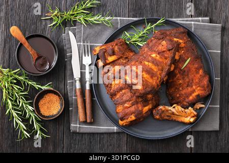 Im Ofen gebackene bbq Baby Schweinerippchen mit frischem Rosmarin auf einem Teller auf dunklem Holztisch mit bbq-Sauce und Besteck, horizontaler Blick von oben, flach gelegt, nicht Stockfoto