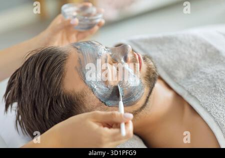 Kosmetologische Hände tragen Ton oder natürliche Schlammmaske auf das Gesicht des Menschen für die Hautreinigung, Verjüngung Stockfoto
