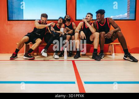 Junge Basketballmannschaft sitzt auf einer Bank in einem Fitnessstudio, lacht und verbindet sich nach einem Spiel, drückt Teamarbeit, Freundschaft und Begeisterung aus. Stockfoto