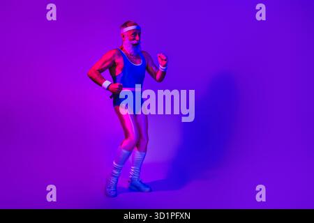 Energiegeladener älterer Mann in Vintage-blauem sportlichem Outfit posiert unter lila Neonlichtern und zeigt Fitness-Freude und Retro-Stil für Werbe-Lifestyle Stockfoto