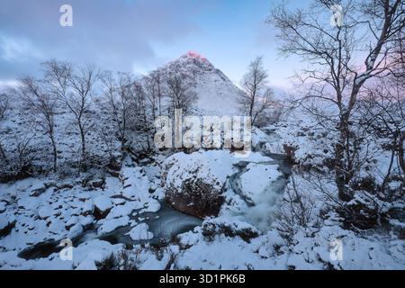 Winterblick auf den Berg Buachaille Etive Mor in Glencoe, schottische Highlands. Schneebedeckte Landschaft mit gefrorenem Fluss und sanftem rosafarbenem Sonnenaufgangslicht, das den Gipfel beleuchtet Stockfoto