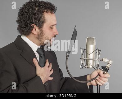 Porträt eines jungen Mannes im Anzug, der mit dem Studio-Mikrofon singt. Isoliert auf grauem Hintergrund. Sängerkonzept Stockfoto