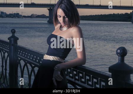 Die Frau wird gegen den Zaun des Dammes gebeugt, steht gegen Wasser und fällt dunkel am Himmel, dramatischer Blick, Frau am Rand, ungewöhnliches Bild, Stockfoto