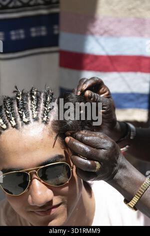 Cordoba, Cordoba - Spanien - 04-08-2024: Junger Mann mit Sonnenbrille, der in Essaouira Zöpfe von erfahrenen Händen bekommt Stockfoto