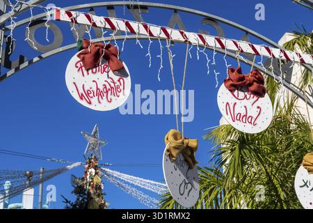 Denia, Alicante - Spanien - 22.12.2023: Hellblauer Himmel mit „Merry Christmas“-Schild in verschiedenen Sprachen und rotem Bogen Stockfoto