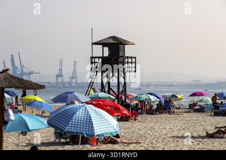 Palmones, Cadiz - Spanien - 25.08.2024: Am Strand steht ein Wachturm mit bunten Sonnenschirmen. Ein Frachter Alamoon segelt auf ruhiger See, ein anderes Schiff Stockfoto
