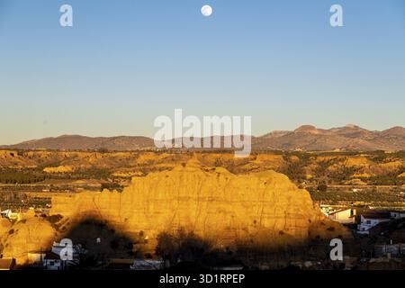 Guadix, Granada - Spanien - 24.01.2024: Ein weitläufiger Blick auf eine Felsformation in Guadix, beleuchtet von der untergehenden Sonne, mit dem Mond im blauen Himmel Stockfoto