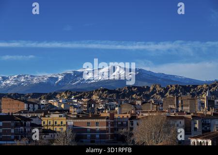 Guadix, Granada - Spanien - 01-24-2024: Das Stadtbild von Guadix zeigt städtische Gebäude im Vordergrund, eingerahmt von einer zerklüfteten Felsenlandschaft und Schnee- Stockfoto