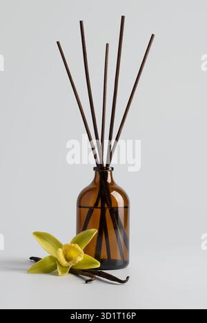 Reed-Diffusor mit Vanillestäbchen in einer braunen Glasflasche und Vanilleblüte auf hellem Hintergrund. Minimale Komposition symbolisiert Ruhe, Reinheit, an Stockfoto