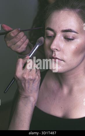 Blauäugige Brünette-Modell genießt professionelles Make-up, Kosmetikerin verwendet weichen Pinsel für Creme Stockfoto