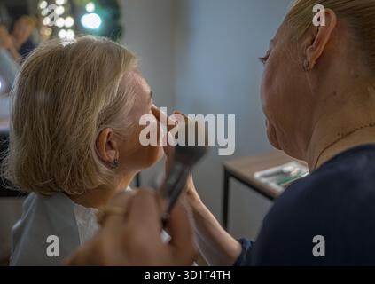 Ein Make-up-Künstler trägt sanft pflegende Creme auf das Gesicht einer blonden Frau mittleren Alters mit einem weichen Pinsel in einem gemütlichen Schönheitssalon auf Stockfoto