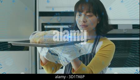 Asiatische Frau, die Tablett mit Brot hält, Ofenhandschuhe in der Küche mit Ofen trägt, mit digitalem Gitter Stockfoto