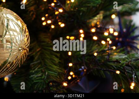 Goldene Kugel mit raffiniertem Glitzerdesign hängt an einem Weihnachtsbaum. Im Hintergrund sind unscharfe grüne Kiefernnadeln und kleine warmweiße Feen-LIG Stockfoto