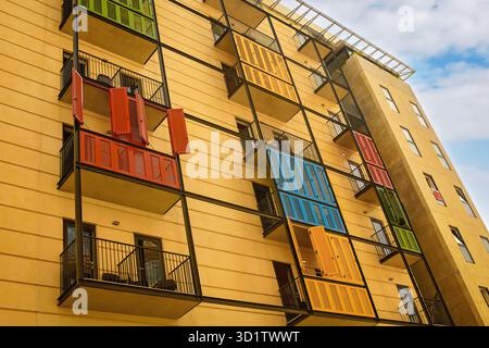 Flacher Blick auf eine helle gelbe, moderne Apartmentfassade mit schwarzen Metallbalkonen und bunten Holzläden unter einem klaren, bewölkten blauen Himmel. Stockfoto