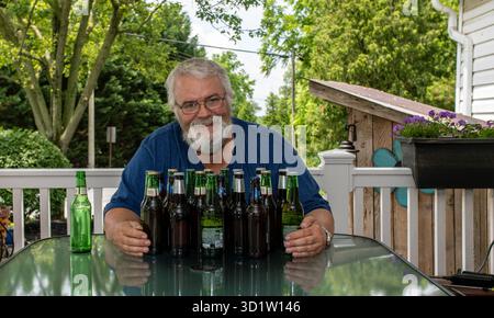 Ein fröhlicher Seniorenmann sitzt an einem Tisch auf einer sonnigen Terrasse, umgeben von mehreren Flaschen Bier. Er lächelt, während er an einem warmen Sommertag mit seiner Kollektion posiert und die Natur genießt. Stockfoto