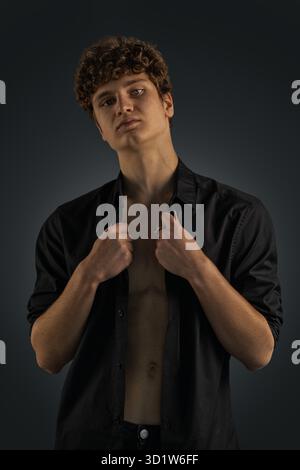 Der junge Mann zeigt einen selbstbewussten Ausdruck, während er sein schwarzes Hemd anzieht. Die Studio-Beleuchtung hebt seine Merkmale hervor und schafft einen markanten Kontrast zum dunklen Hintergrund. Stockfoto