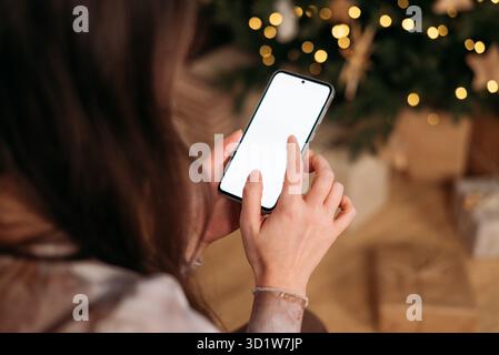 Die Hände einer Frau mit einem vertikalen Smartphone mit einem leeren weißen Bildschirm, einem verschwommenen Weihnachtsbaum und Geschenken im warmen Hintergrund. Stockfoto