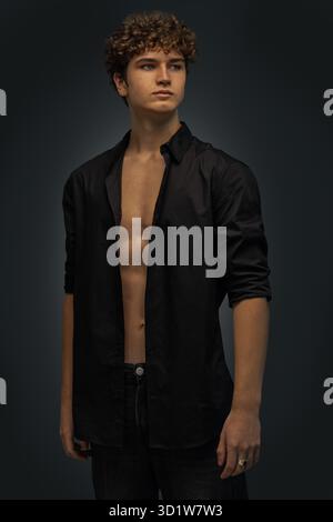 Ein junger Mann steht in einem Studio und trägt ein schwarzes Hemd mit lockigen Haaren. Er zeigt eine selbstbewusste Haltung, die das Wesen des modernen Stils und der Jugend einfängt. Stockfoto