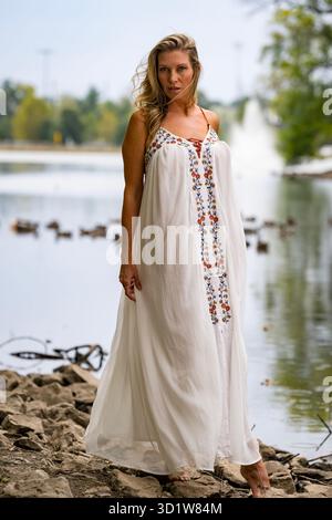 Das blonde Modell strahlt Eleganz aus in White Dress am Serene Lake, gebadet in der Spätsommersonne Stockfoto