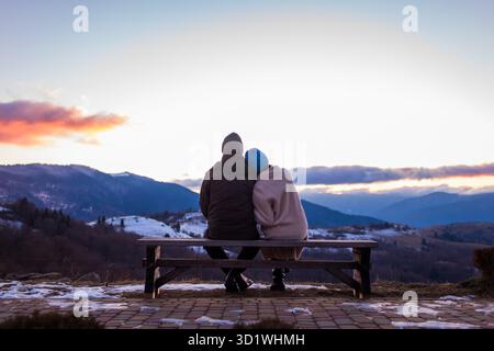 Verliebte Paare genießen die Landschaft der Winterkarpaten, die bei Sonnenuntergang auf einer Bank sitzen. Urlaub. Rückansicht. Leerraum Stockfoto