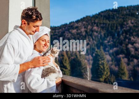 Junges Paar, das eine Katze auf dem Hotelbalkon hält und die Winterberglandschaft im Bademantel genießt. Reisen Sie mit Haustier. Mann und Frau, die mit DO spielen Stockfoto