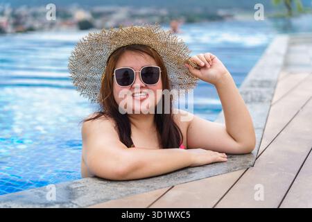 Übergewichtige junge asiatische Frau trägt orange Badeanzüge und Strohhut am Strand, die sich im Pool entspannt, glückliche und große Frau fröhlich lustig Stockfoto