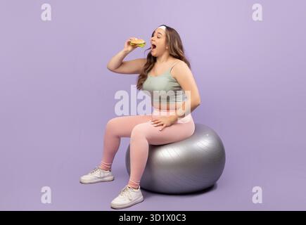 Fröhliche junge Frau, die einen Burger genießt, während sie auf einem Fitnessball in einem Studio balanciert Stockfoto