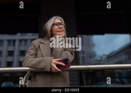 Ältere Geschäftsfrau denkt über Entscheidungen nach, während sie ein Smartphone in der Nähe ihres Büros hält, in einer städtischen Umgebung während der Tageslichtstunden Stockfoto