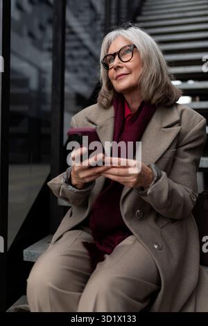 Ältere Frau, die tagsüber auf einer Treppe in einem modernen Gebäude sitzt und ein Smartphone benutzt, während sie in stilvoller Winterkleidung gekleidet ist Stockfoto