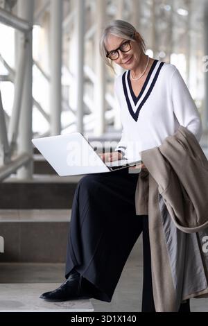 Geschäftsfrau mittleren Alters, die nachmittags in einer modernen Büroumgebung drinnen sitzt und an einem Laptop arbeitet Stockfoto