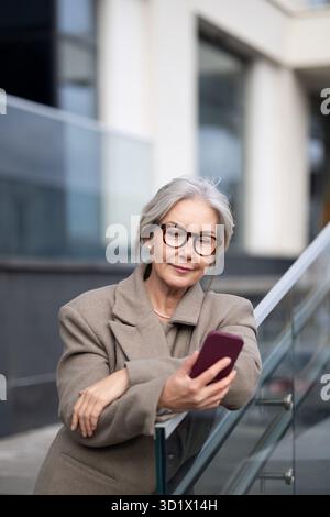 Eine ältere Geschäftsfrau, die ihr Telefon überprüft, während sie nachmittags vor einem modernen Bürogebäude steht Stockfoto