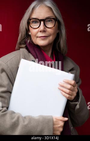 Eine kaukasische Geschäftsfrau mittleren Alters hält einen Laptop und posiert selbstbewusst vor einem leuchtend roten Hintergrund in einer urbanen Umgebung Stockfoto