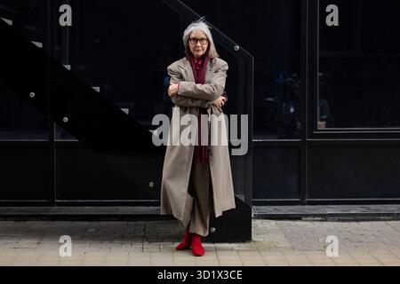 Stilvolle ältere Frau in einem langen Mantel und roten Stiefeln steht an einem sonnigen Nachmittag selbstbewusst vor einer modernen städtischen Kulisse Stockfoto