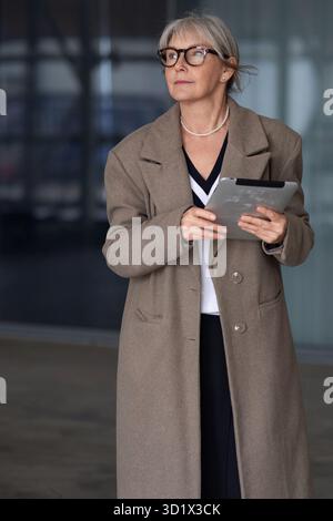 Eine ältere Frau in Brille und braunem Mantel, die eine Tafel hält, während sie an einem windigen Tag vor einem modernen Gebäude steht Stockfoto