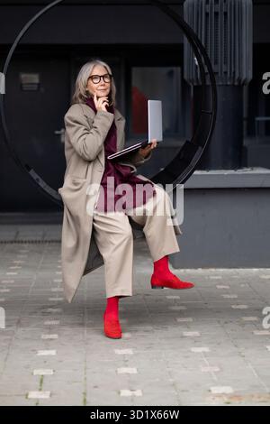 Stilvolle ältere Frau, die nachmittags im Kreis sitzt, während sie ihren Laptop in einer modernen städtischen Umgebung benutzt Stockfoto