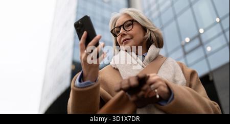 Eine reife Frau, die ihr Smartphone überprüft, während sie an einem bewölkten Tag draußen in einem Stadtgebiet steht Stockfoto