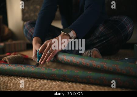 Eine Person im karierten Pyjama schmückt blaue Geschenkverpackung mit Blumenmuster in einem gemütlichen Zuhause, mitten im Projekt, und hält eine warme, raffinierte Szene der Geschenkvorbereitung fest Stockfoto