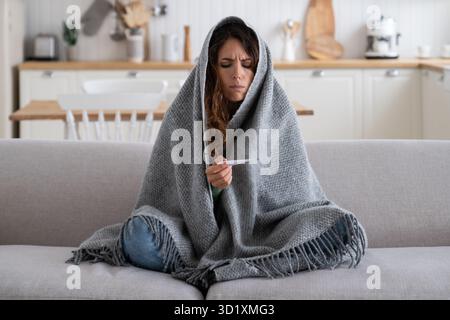 Eine kranke, traurige Frau, die in eine Decke gewickelt ist und thermomete ansieht, sitzt auf dem Sofa in einer unbeheizten Wohnung Stockfoto