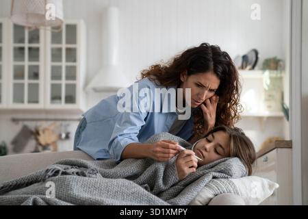 Besorgte Mutter, die Thermometer ansieht und die Temperatur der kranken Tochter im Bett prüft Stockfoto