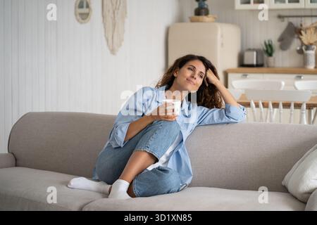 Junge glückliche, entspannte Frau, die auf dem Sofa sitzt und zu Hause ruhige Momente mit einer Tasse Tee oder Kaffee genießt Stockfoto