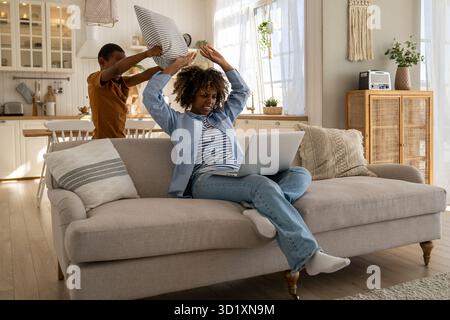 Die wütende mutter putzt sich mit der Hand ab, Sohn spielt mit Kissen und lenkt von der Arbeit auf der Couch mit Laptop ab Stockfoto