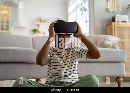 Ein besessener schwarzer Junge, der virtuelle 3D-Realität in einer VR-Brille erlebt, sitzt zu Hause in der Gadget-sucht Stockfoto