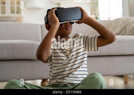 Ein besessener schwarzer Junge, der virtuelle 3D-Realität in einer VR-Brille erlebt, sitzt zu Hause in der Gadget-sucht Stockfoto