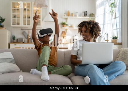 Ein Junge, der in virtueller Realität spielt, während er neben seiner mutter auf dem Sofa sitzt und an einem Laptop arbeitet Stockfoto