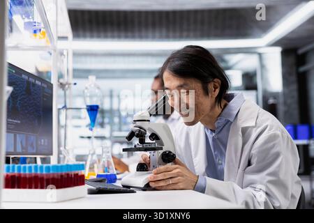 Lächelnder Mikroskopist mit Mikroskop, zufrieden mit detaillierten Ergebnissen der Probenbeobachtung in kontrollierter Laborumgebung. Glücklicher Biologe zufrieden mit dem, was er im Gewebe unter dem optischen Gerät sieht Stockfoto